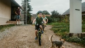 Two kids cycling on a dirt path with a small dog chasing them, adult supervising nearby.